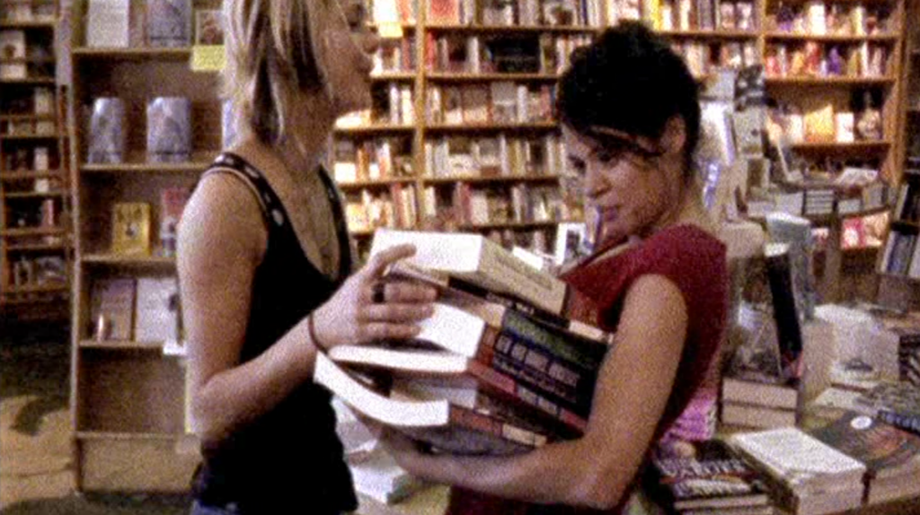 Sadie (played by Nicole Vicius) piles another book on the stack in Anna's arms in the bookstore.