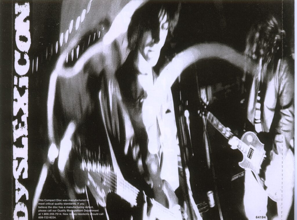 black and white photograph of the band playing, only the two guitar players really appearing in frame; "Dyslexicon" along the left side as to appear even when CD cover is closed on the spine.