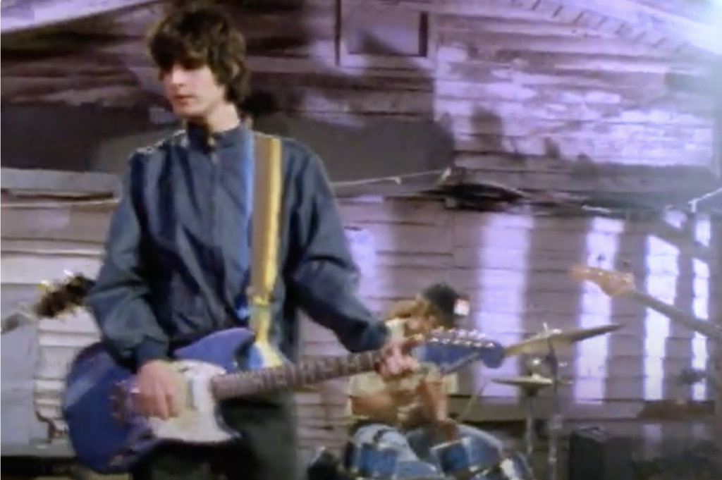 still from "Weird Out" music video of Kevin playing guitar, Dante in background on drums