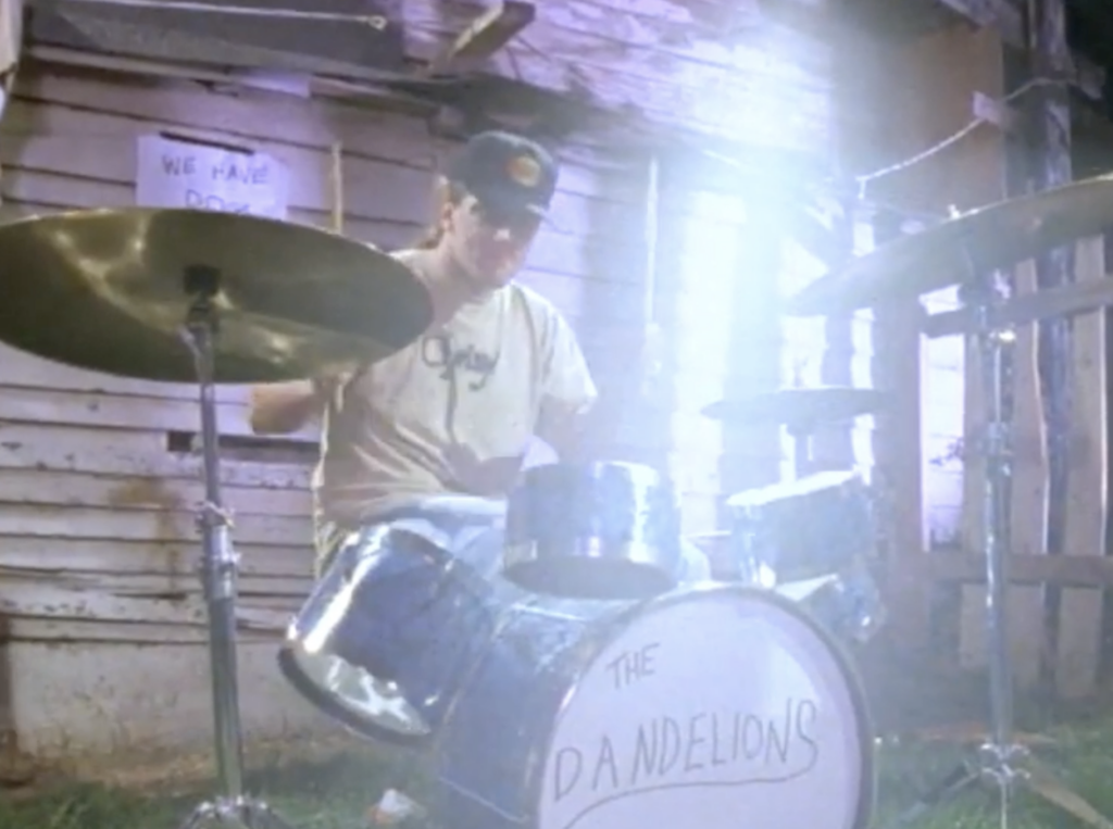 still from "Weird Out" music video of Dante playing small drum kit, "The Dandelions" written on his kick drum